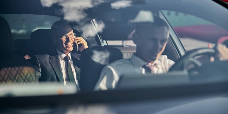 Man is driving comfortable car while his boss is speaking on cell phone in back